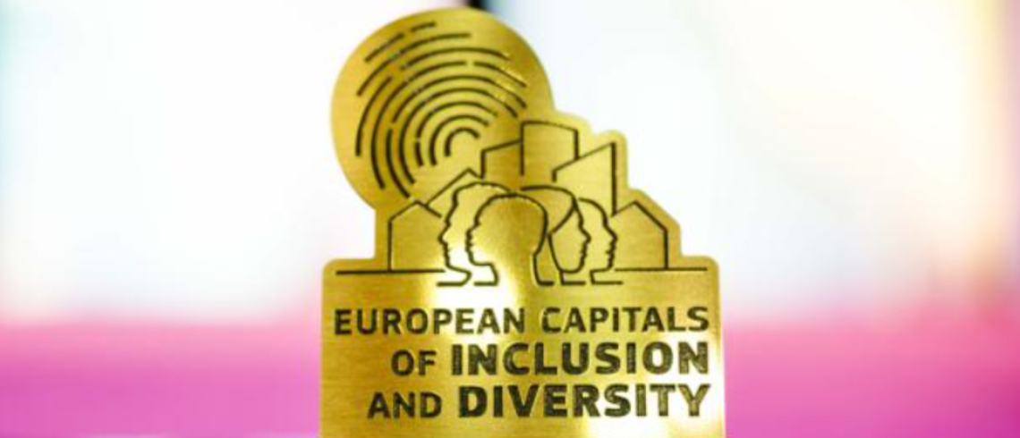 L’image montre un trophée doré, posé sur un socle noir, portant l'inscription “European Capitals of Inclusion and Diversity”. La silhouette du trophée est stylisée pour refléter son symbole : en arrière-plan, on distingue des bâtiments urbains, accompagnés d’un motif circulaire rayonnant, évoquant une empreinte. Au premier plan, un groupe de quatre personnes de profil est visible : deux tournent la tête vers la droite et deux vers la gauche. Le fond est flou, avec des tons clairs et pastel (rose, blanc, vio